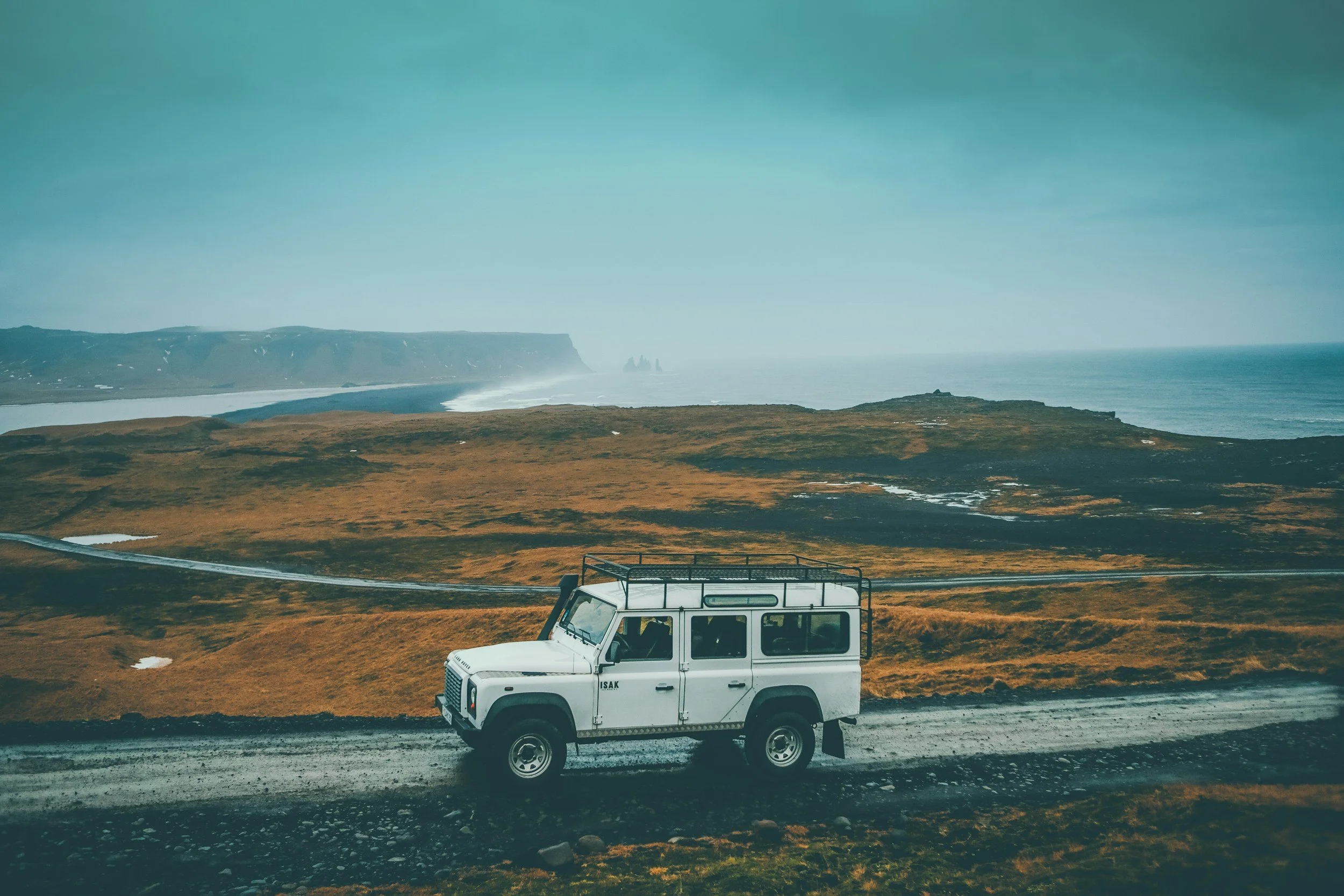 A white off-road vehicle driving on a gravel road through a remote, grassy landscape with cliffs and the ocean in the background.