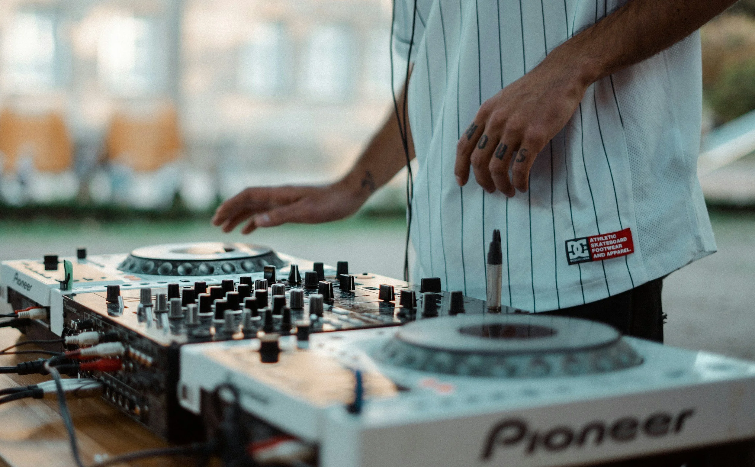 Person DJing using Pioneer turntables outdoors, wearing a light blue striped shirt.