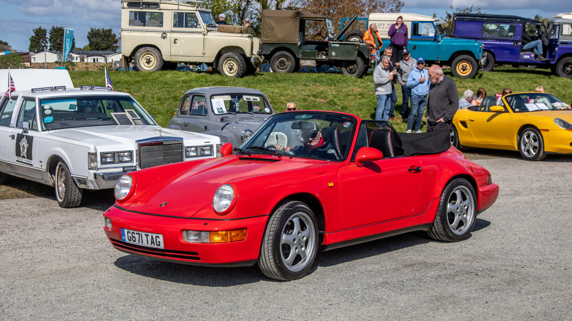 A red Porsche 911 convertible parked among various vintage and classic cars during an outdoor car show. People are walking around and chatting in the background.