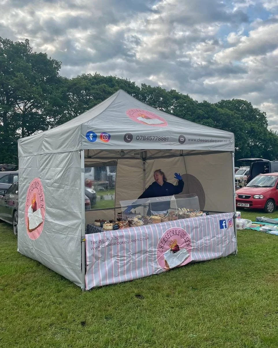 A woman standing behind a cheesecake stand at an outdoor market. The stand has a banner with the logo 'Cheesecake Queen', displaying various cakes. The tent has social media icons and contact information, with parked cars and green trees in the background.
