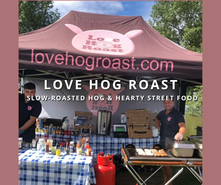 A food stall at an outdoor event selling slow-roasted hog and street food. The stall has a maroon canopy with the words 'lovehogroast.com' and a pig logo. Two men are working behind the stall, which is set up with various cooking equipment, condiments, and a checkered tablecloth.