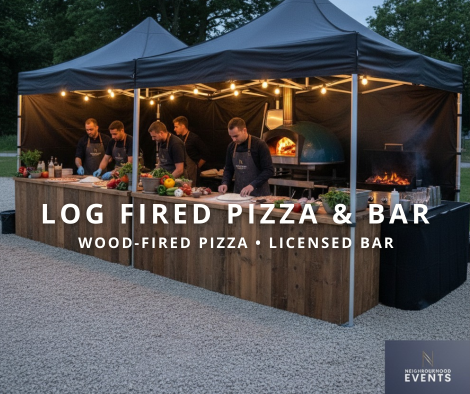 Outdoor setup with a black canopy tent, under which five chefs in black aprons are preparing log-fired pizza. The serving area is a wooden counter with fresh vegetables and food ingredients. There is a wood-fired pizza oven with flames visible inside and a separate grill with fire. String lights are hanging from the canopy. The setting is outdoors with trees in the background.