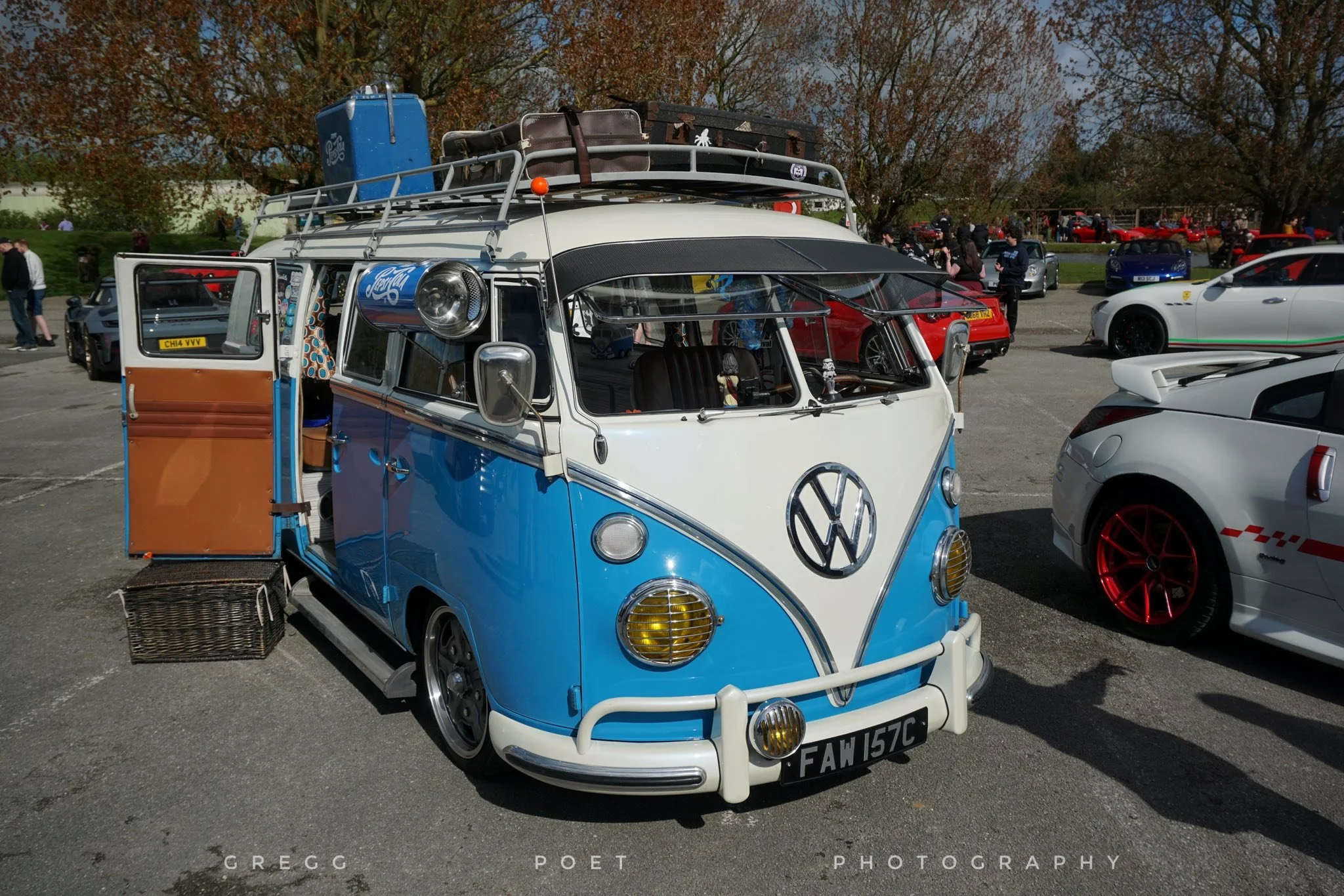 Vintage Volkswagen van painted in blue and white, parked at a car gathering, with luggage and a camera on the roof rack, surrounded by other cars and people.