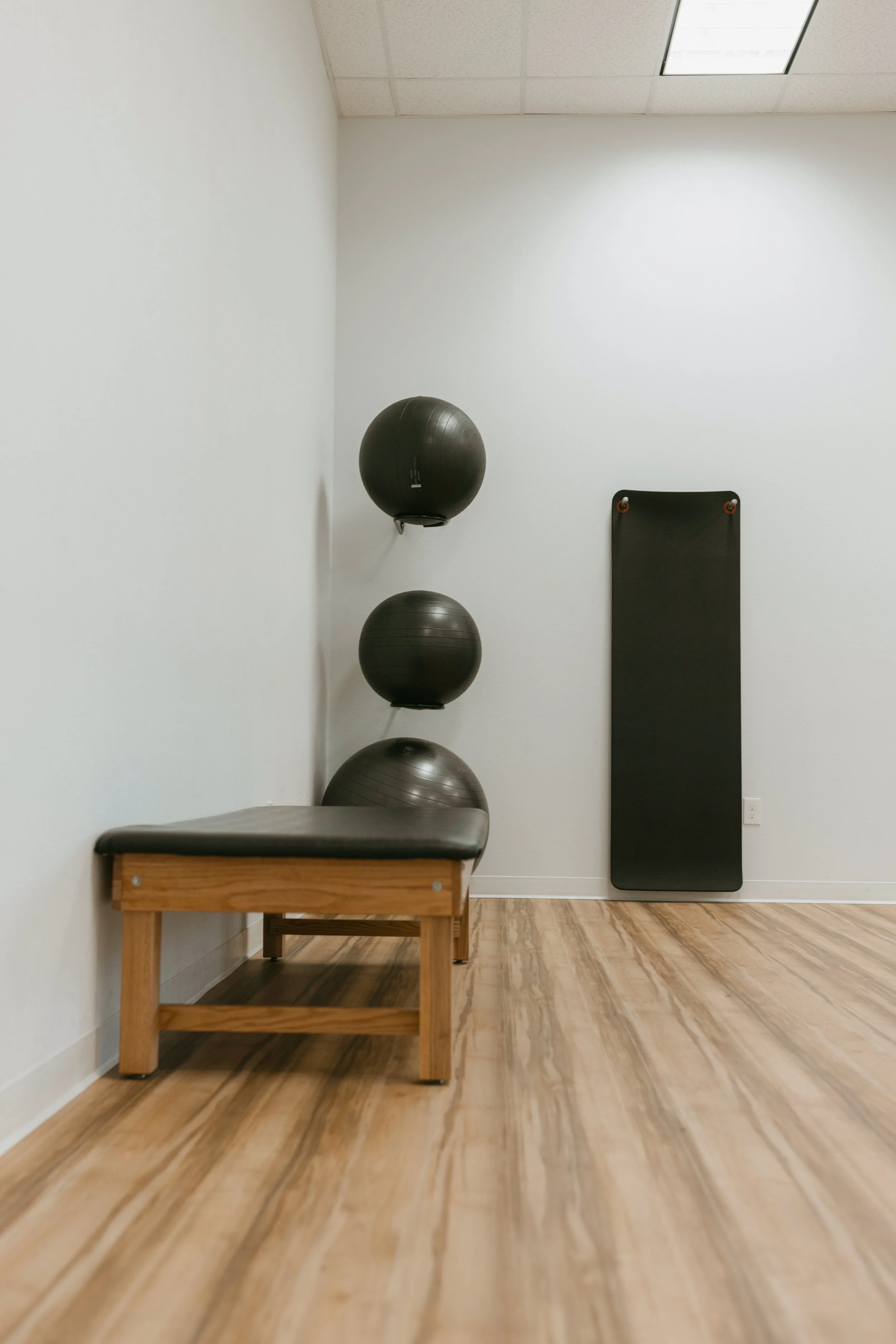 A small gym room with a wooden bench with black padding, three black exercise balls mounted on the wall, a black padded mat hanging vertically, and a ceiling light panel.