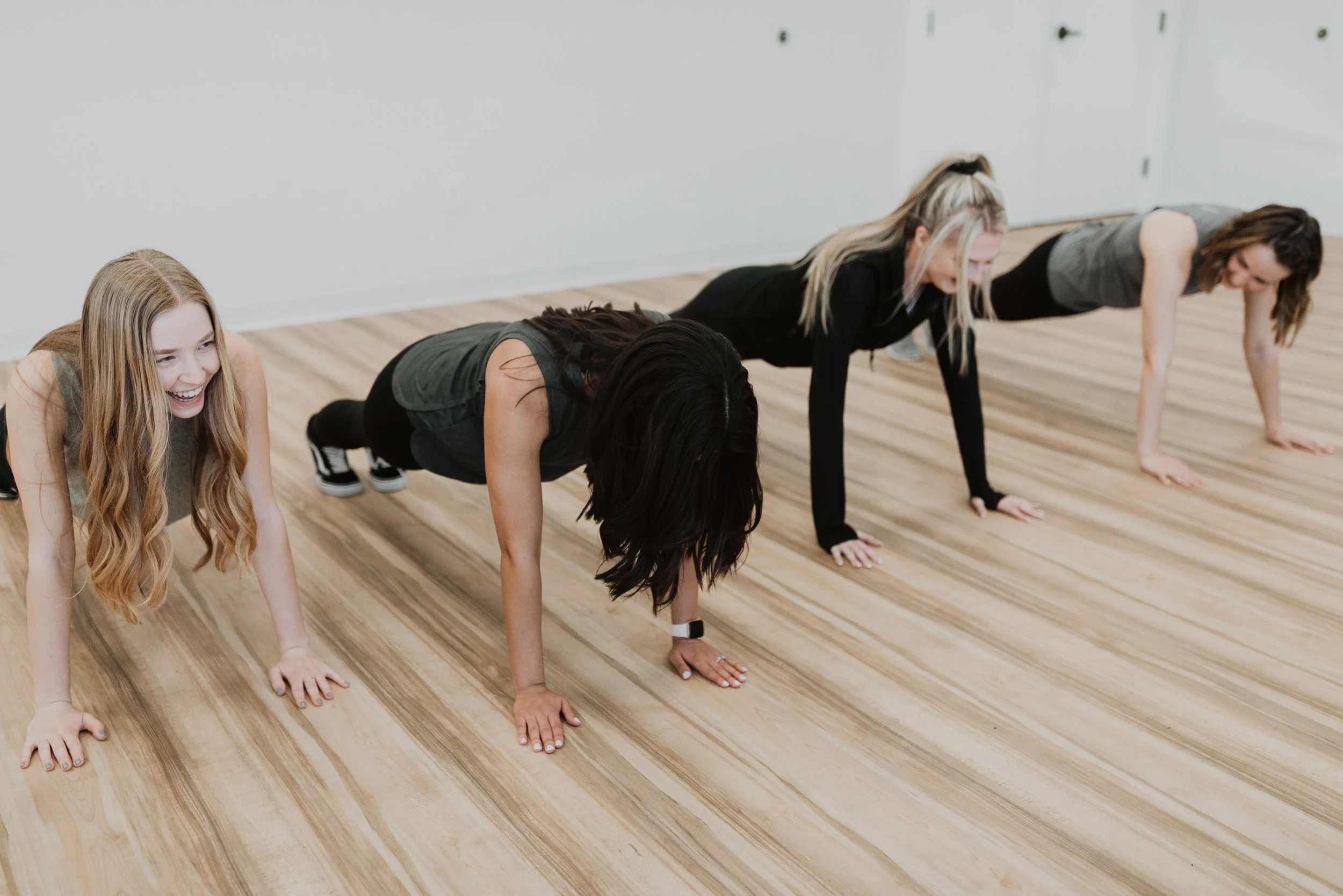 Four women doing push-ups together in a fitness class, smiling and engaging in exercise on a wooden floor.