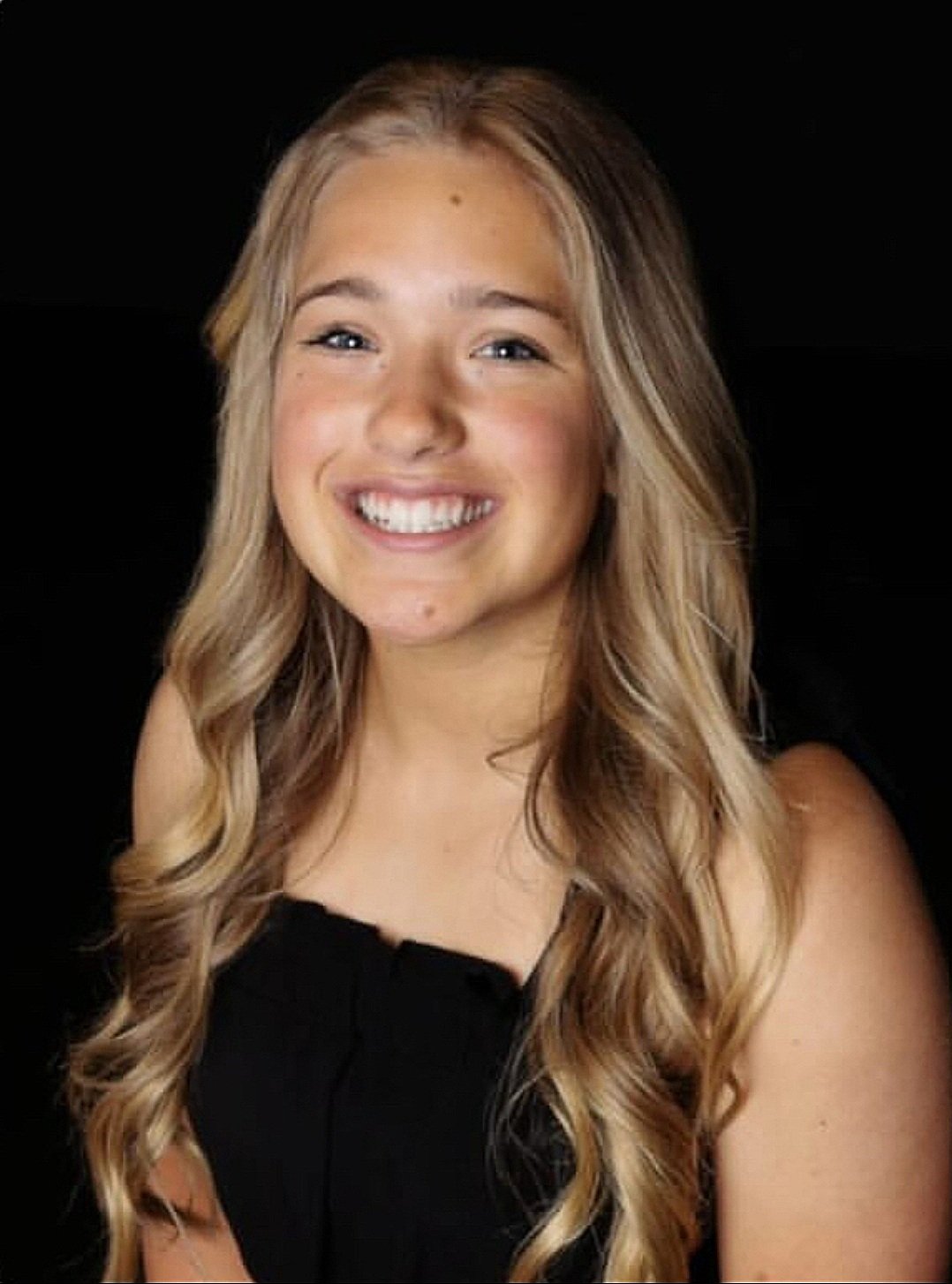 A young woman with long, wavy blonde hair smiling at the camera, wearing a black sleeveless top, against a black background.