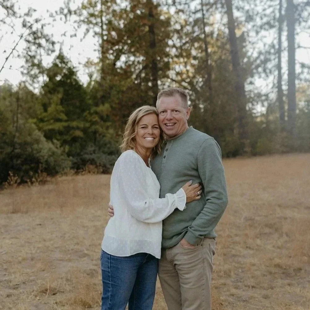 A smiling couple standing outdoors in a grassy field with trees in the background, embracing each other.