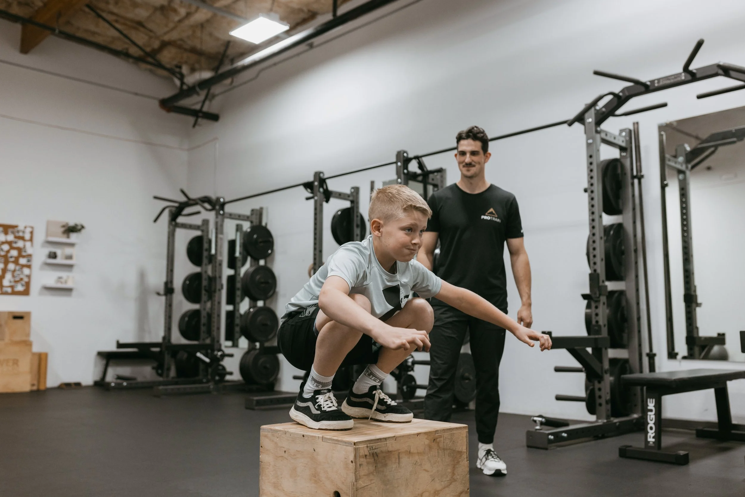 A boy performs a box jump exercise in a gym, with a trainer supervising behind him.