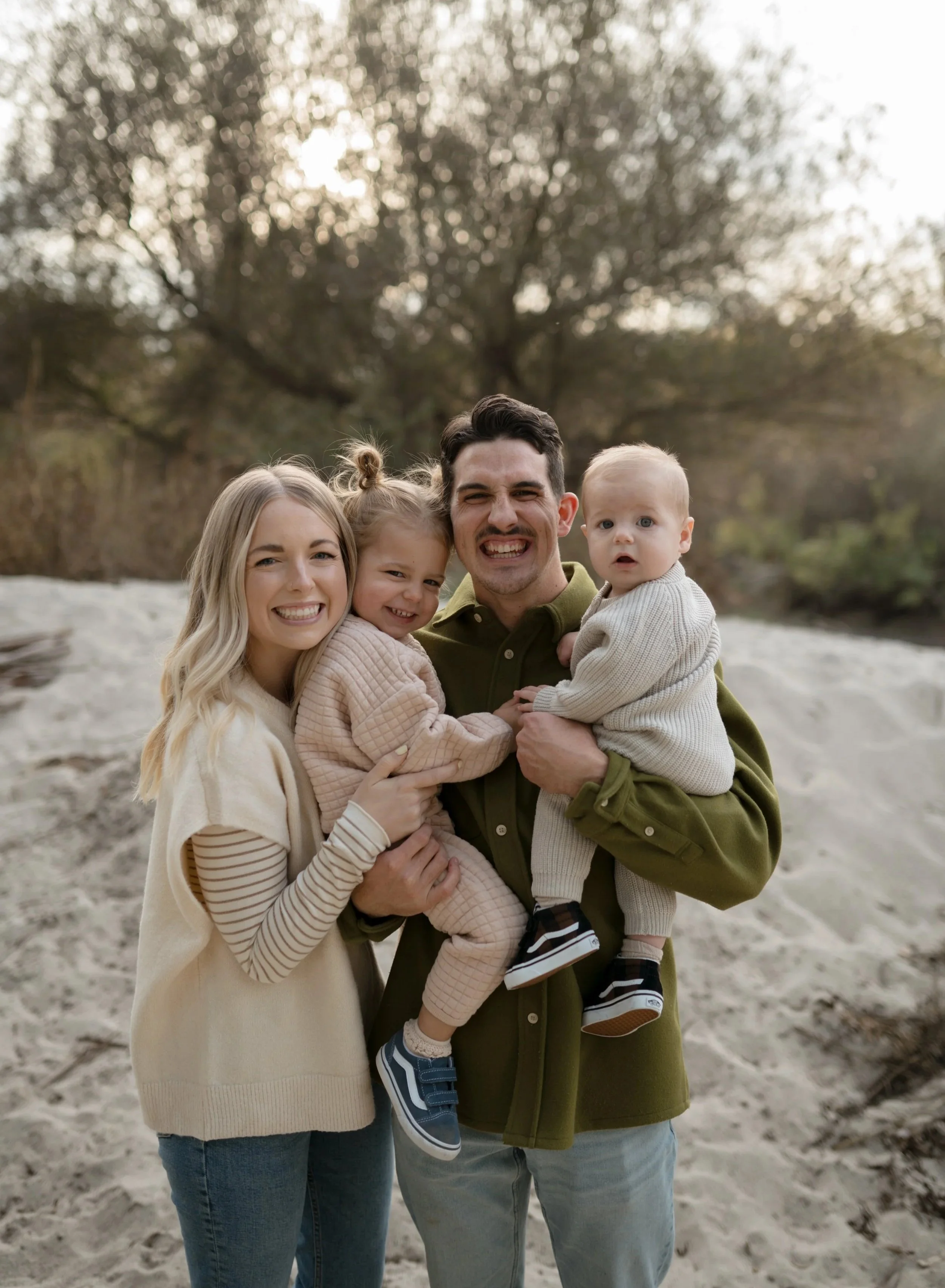 Family of four smiling and posing together outdoors on a sandy area with trees in the background, during what appears to be late afternoon or early evening.