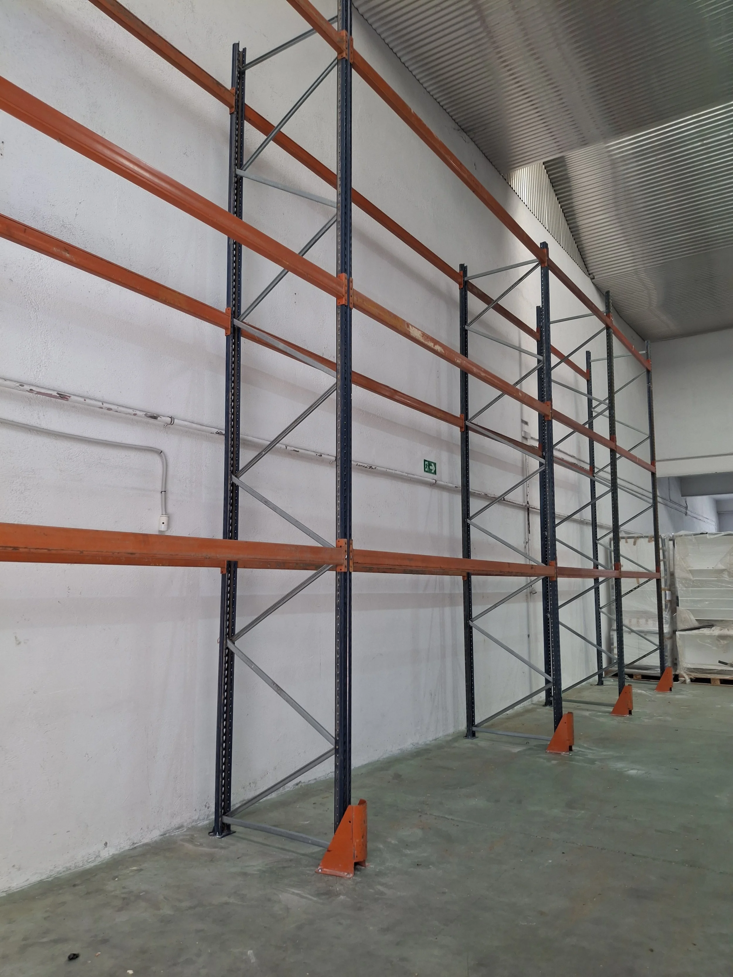 Estanterías metálicas en un almacén. Piso de concreto, paredes blancas y techo de lámina corrugada.