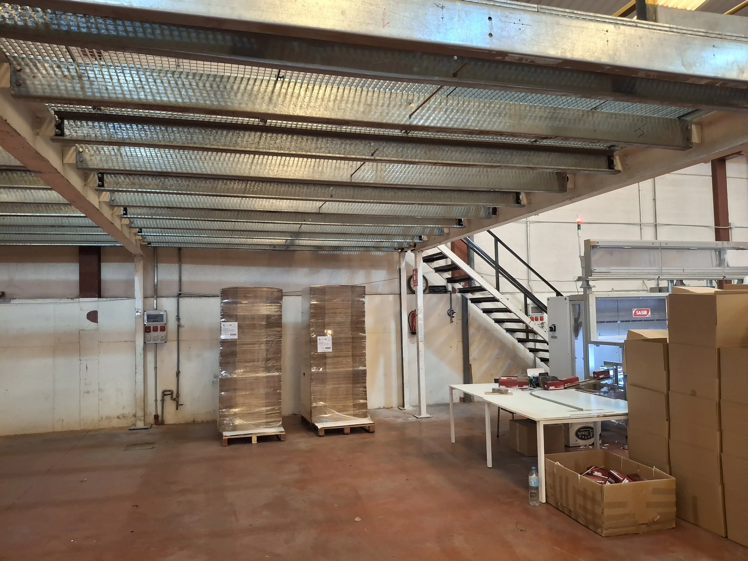 Interior de una fábrica o almacén con estructura de metal y escaleras, mesas de trabajo y cajas de cartón almacenadas, con un ambiente organizado y industrial.