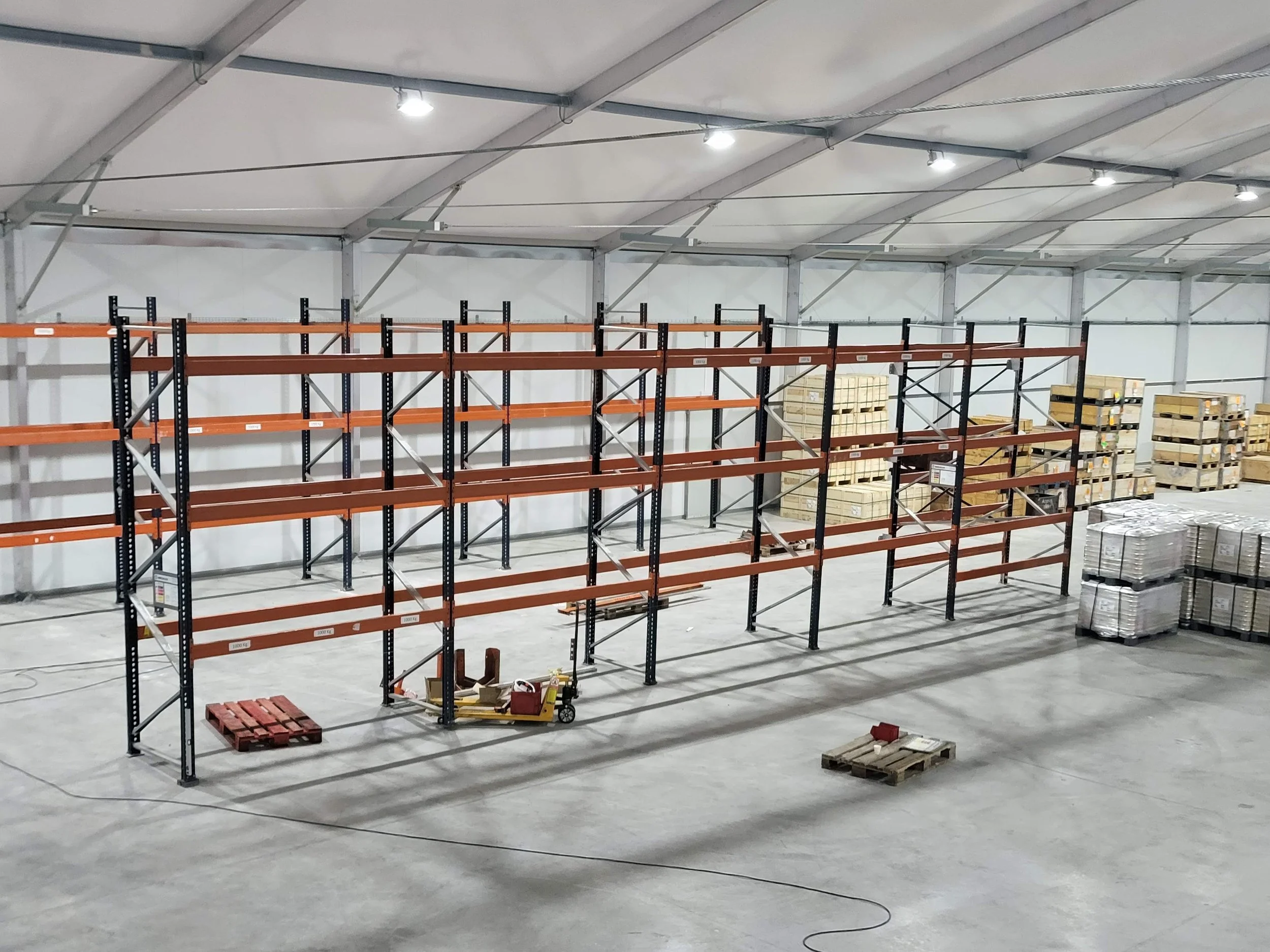 Interior de un almacén en construcción con estanterías vacías, pallets de madera y materiales de construcción en el suelo.