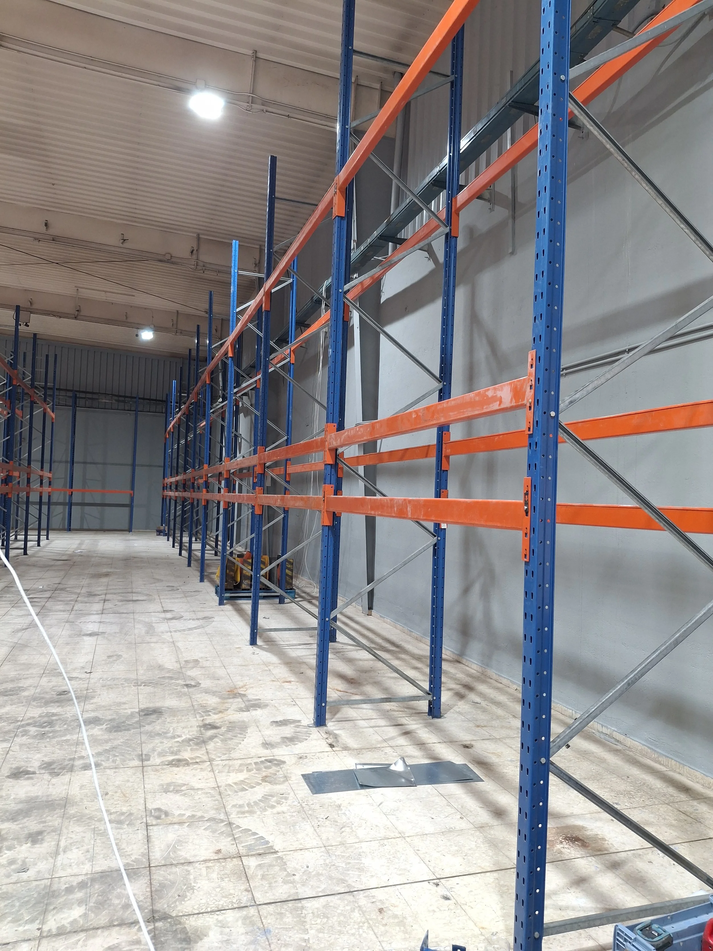 Una estructura de estantería metálica de color azul y naranja en un espacio interior que parece estar en construcción o almacenamiento, con suelo de baldosas y paredes de diferentes colores.