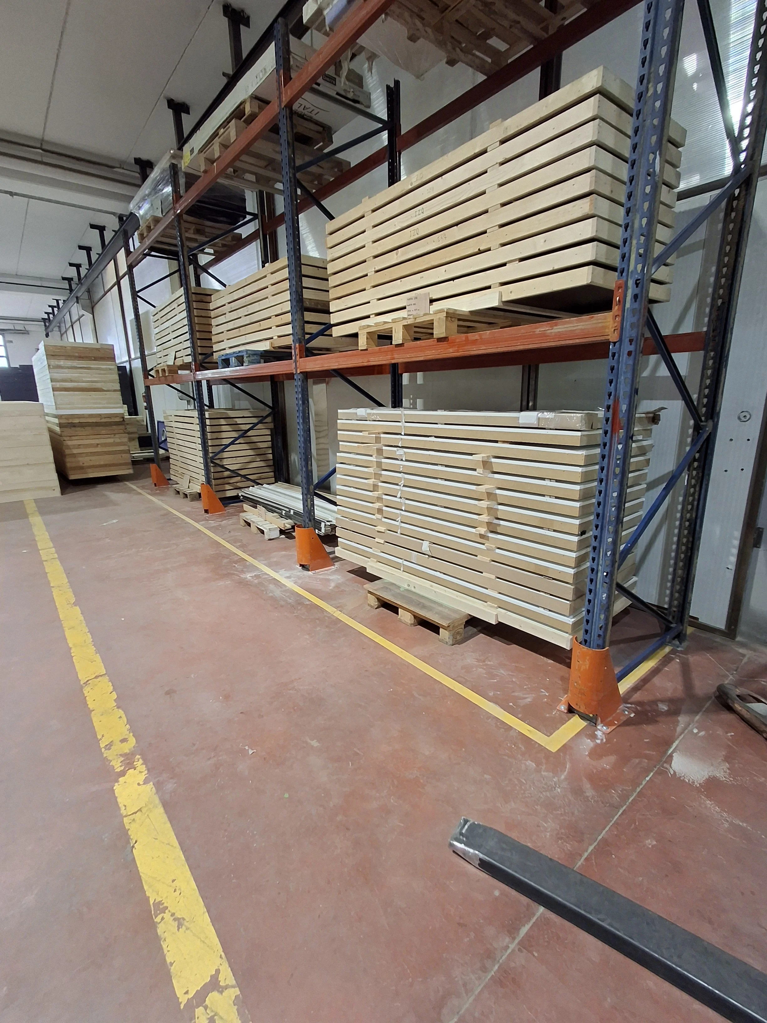 Estantería de metal con varias tablas de madera apiladas en un almacén, con una línea amarilla en el suelo y una estructura de techo en el fondo.