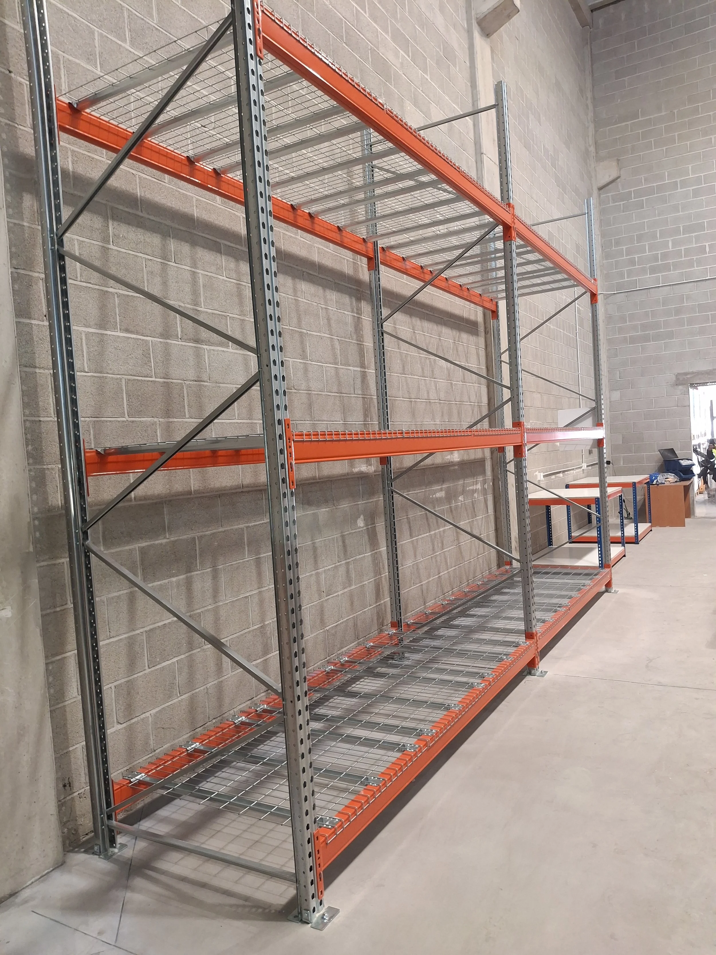 Estanterías vacías en un almacén, hechas de metal con estructura de color gris y estantes de rejilla, ubicadas contra una pared de bloques de cemento.