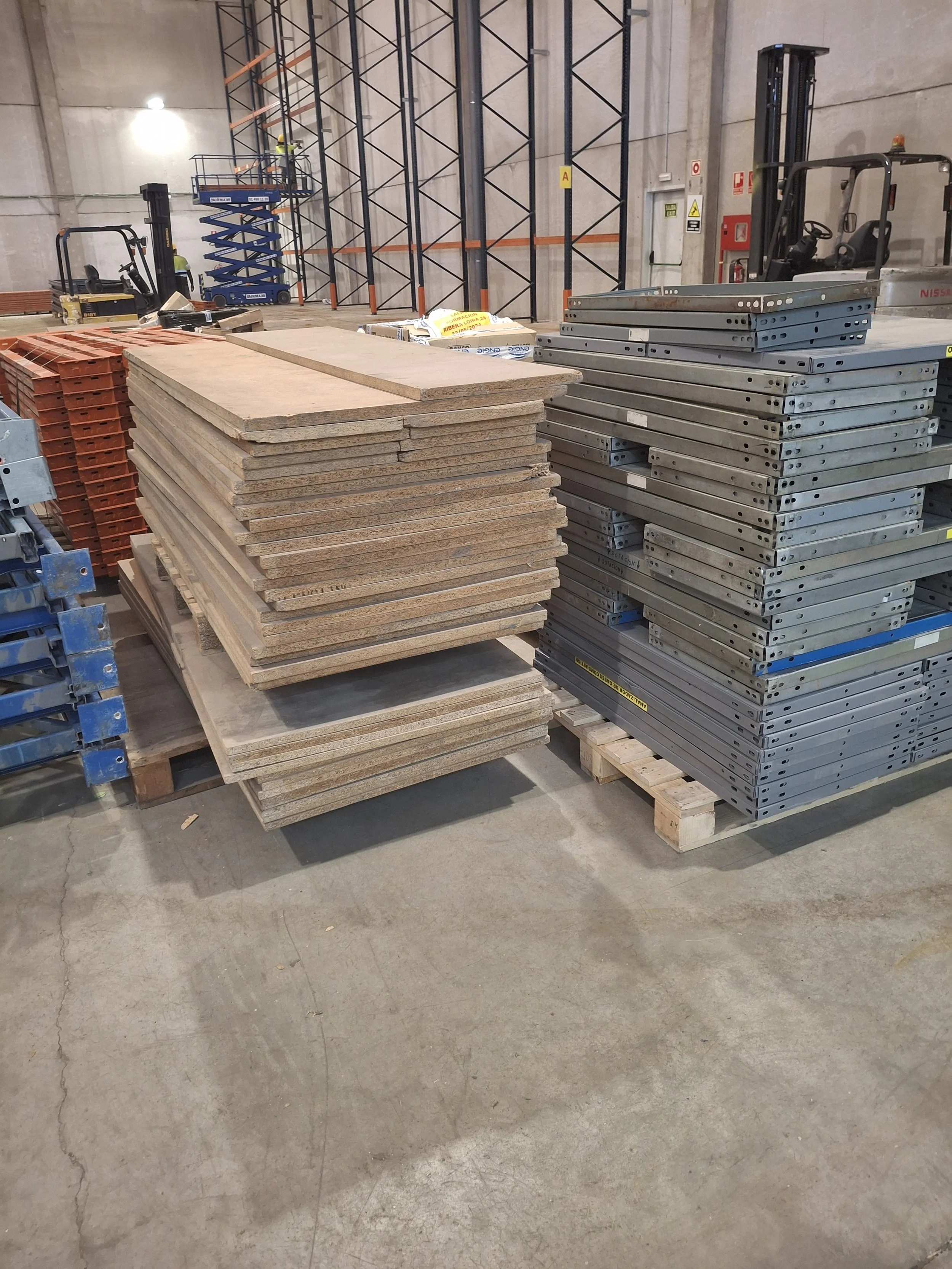Imagen de un almacén con estanterías metálicas y plataformas de madera y metal apiladas, además de unidades de andamios y maquinaria de construcción.
