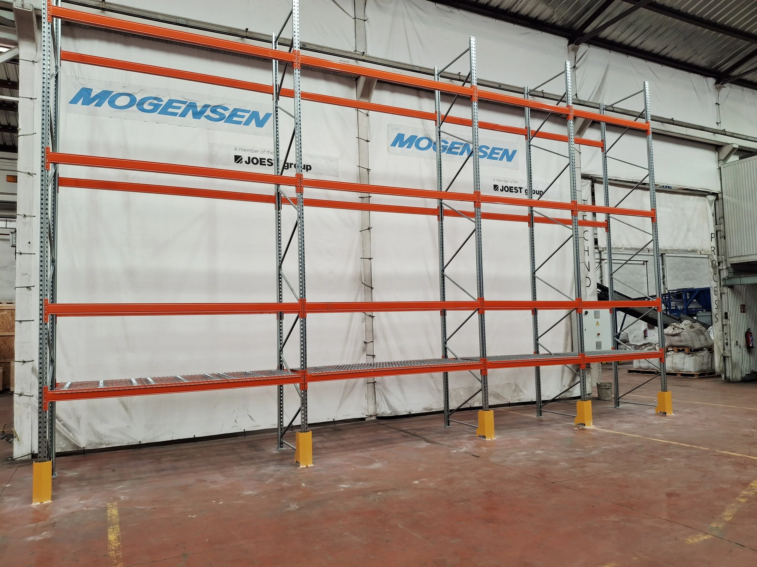 Estanterías de metal ancladas en una estructura de almacenaje dentro de un almacén industrial, con logos de Mogens en la parte superior.