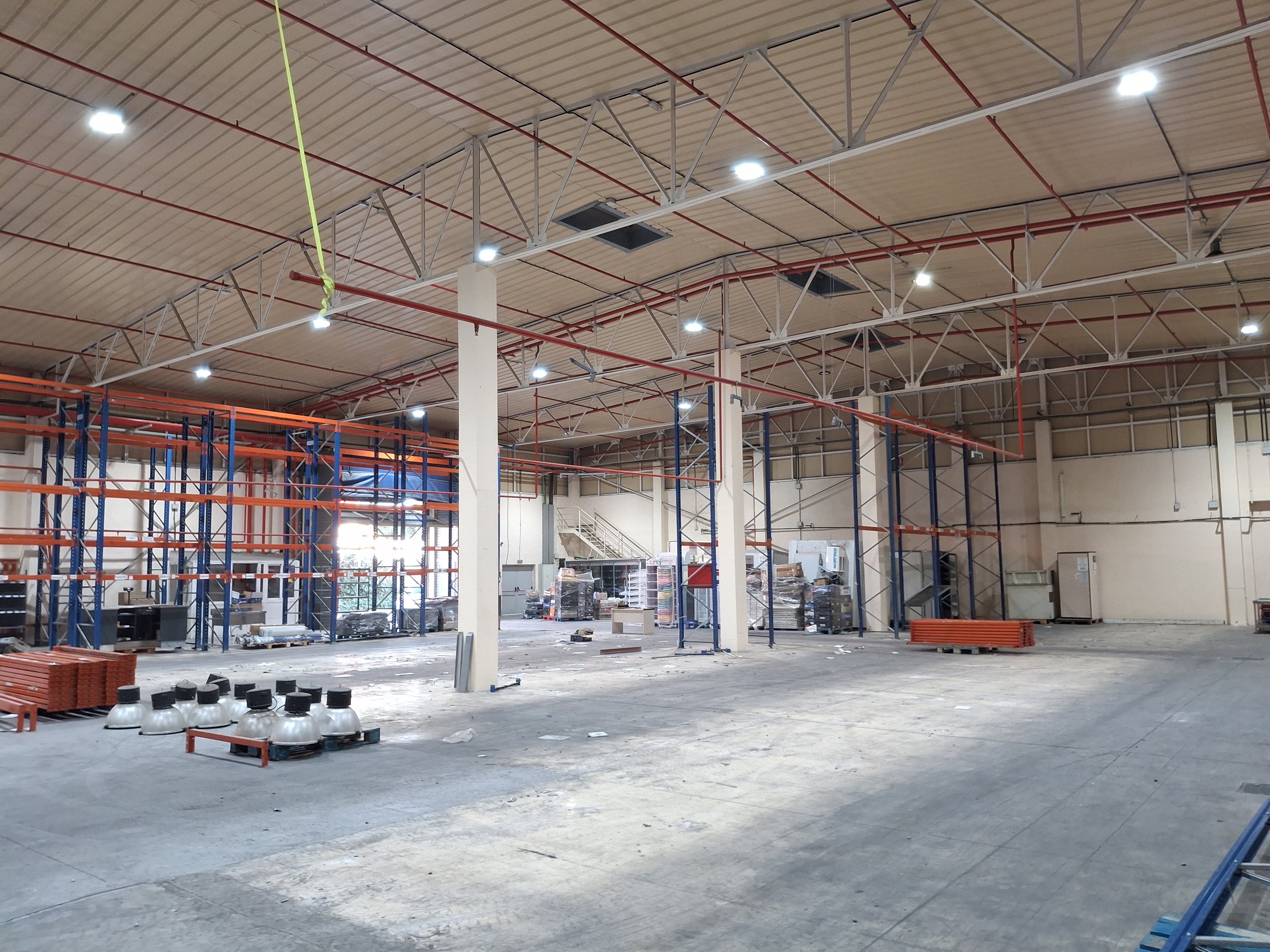 Interiores de un almacén o centro de distribución en construcción, con estanterías metálicas vacías y herramientas en el suelo, techos altos con iluminación moderna y estructura de soporte visible.