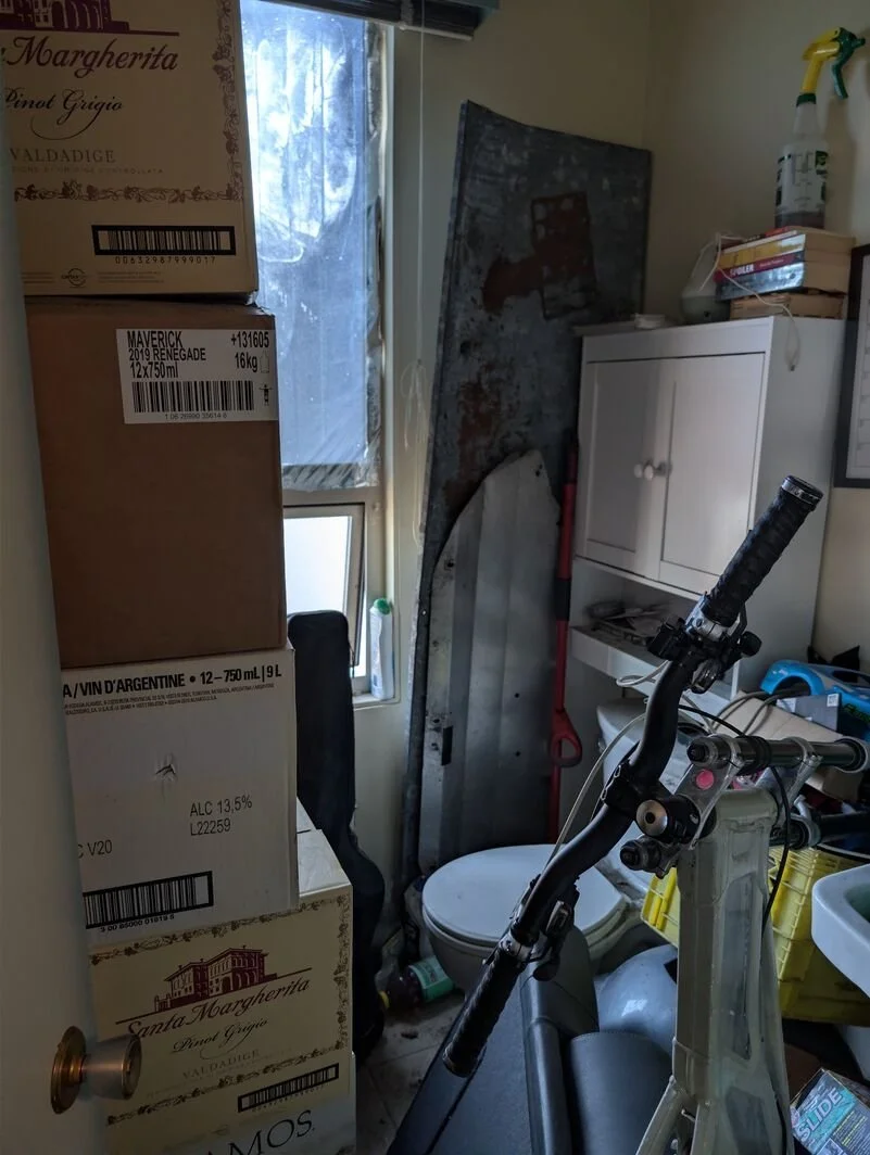 The bathroom is cluttered with stacked boxes, a white cabinet, a bicycle, and various cleaning supplies, with a window showing trees outside.