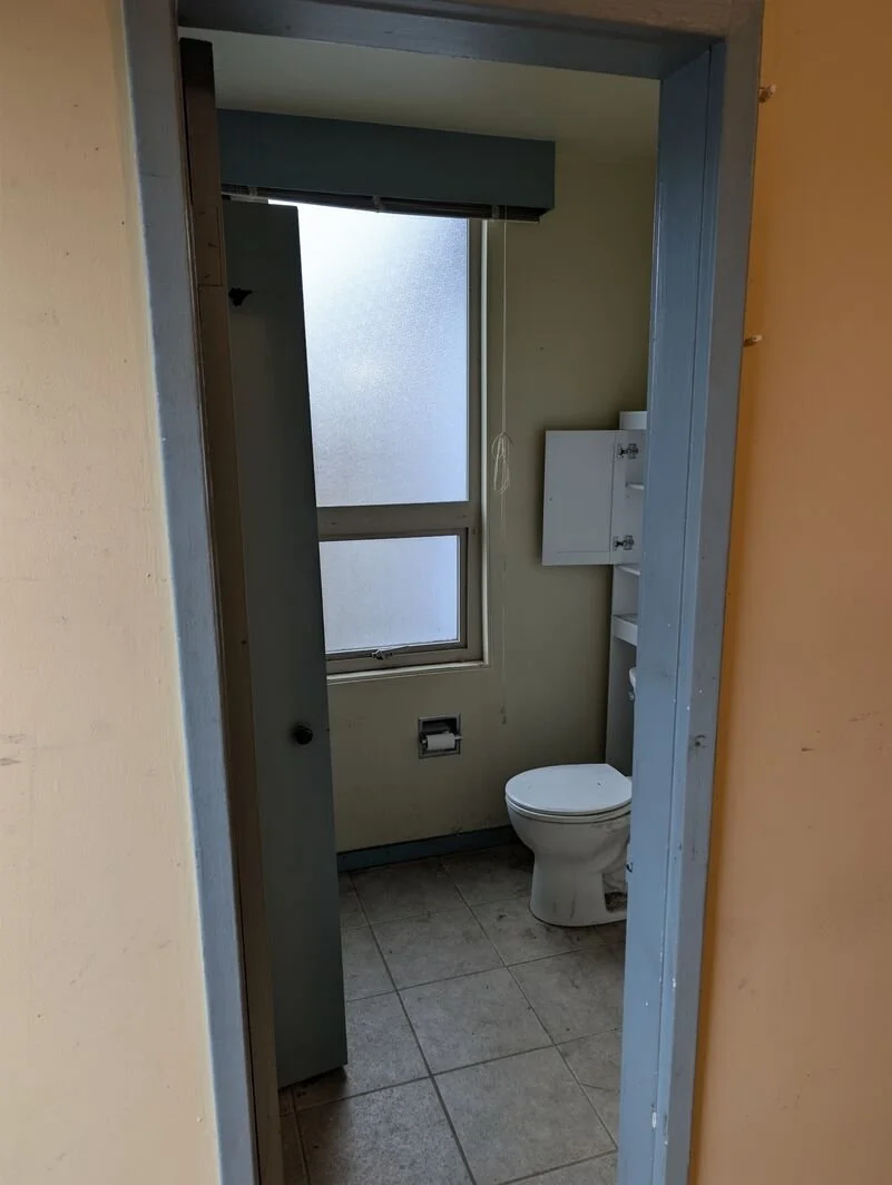 A small, unfinished bathroom with a frosted window, a toilet, a partially open cabinet, and a tiled floor.