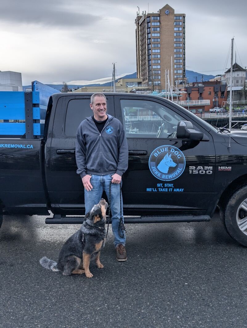Man standing beside a Blue Dog Junk Removal pickup truck while his dog sits looking up at him near a waterfront marina.