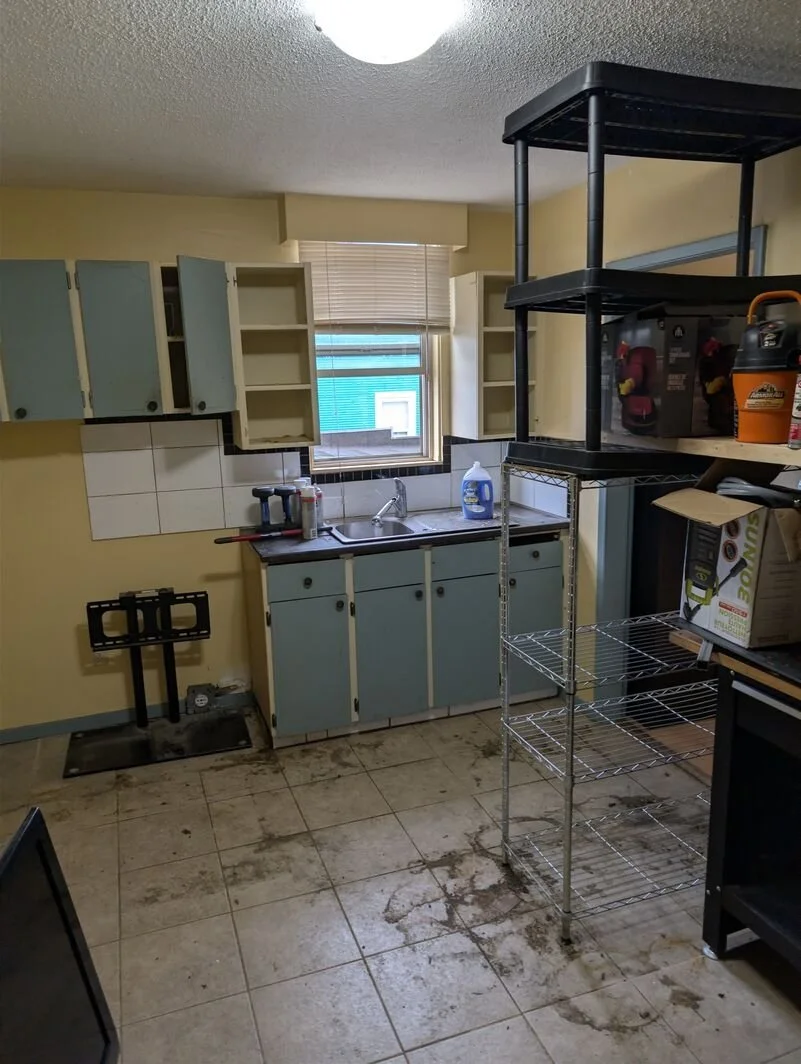 Kitchen with dirt and debris on the tiled floor, open cabinets, a small window with blinds, a metal shelving unit, and various cleaning supplies.