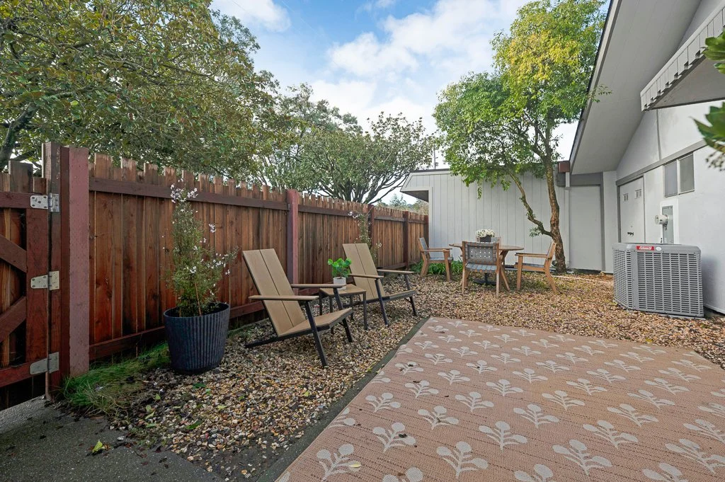 Fenced backyard with outdoor furniture, including chairs, a table with flowers, and a patterned rug, surrounded by trees and a house with an outdoor air conditioning unit.
