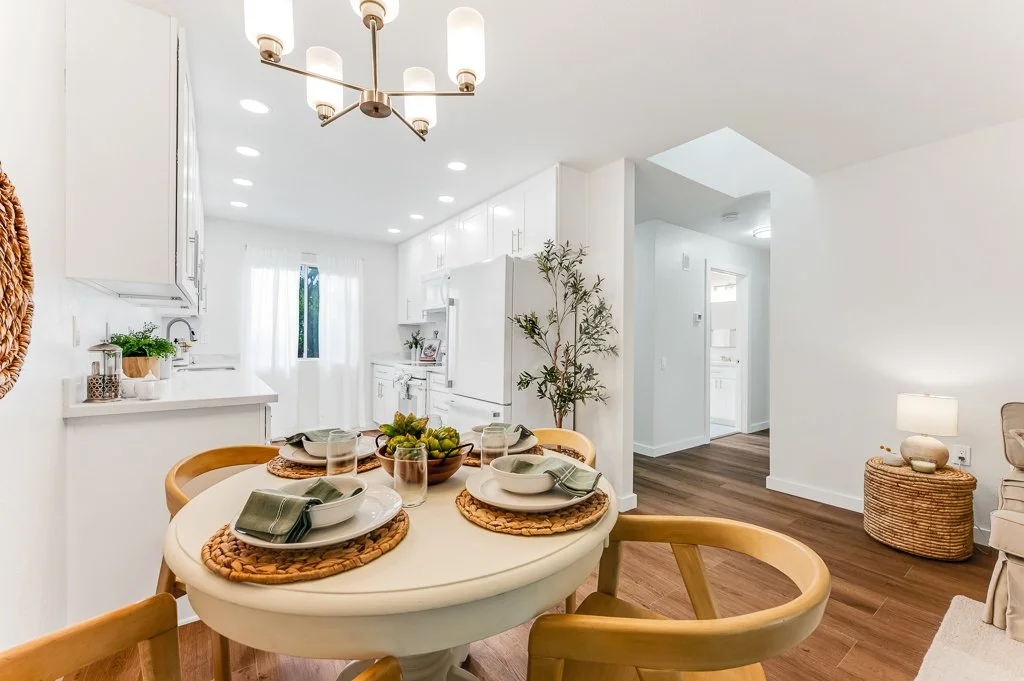 A dining area with a white round table set with plates, glasses, and napkins, surrounded by wooden chairs, in a bright, modern kitchen with white cabinets, a chandelier, and hardwood flooring.