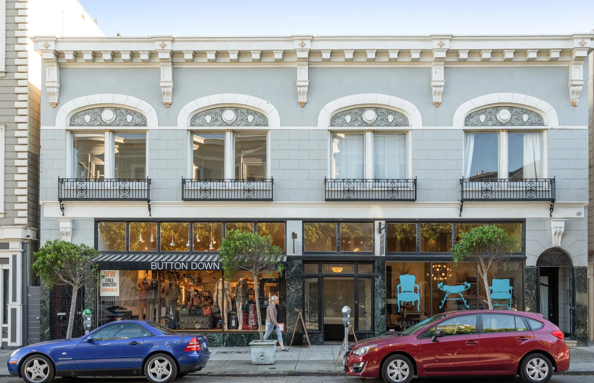 Gray three-story building with large front windows and decorative white trim, ground floor retail store with sign 'Button Down', cars parked in front, trees lining sidewalk, person walking on sidewalk.