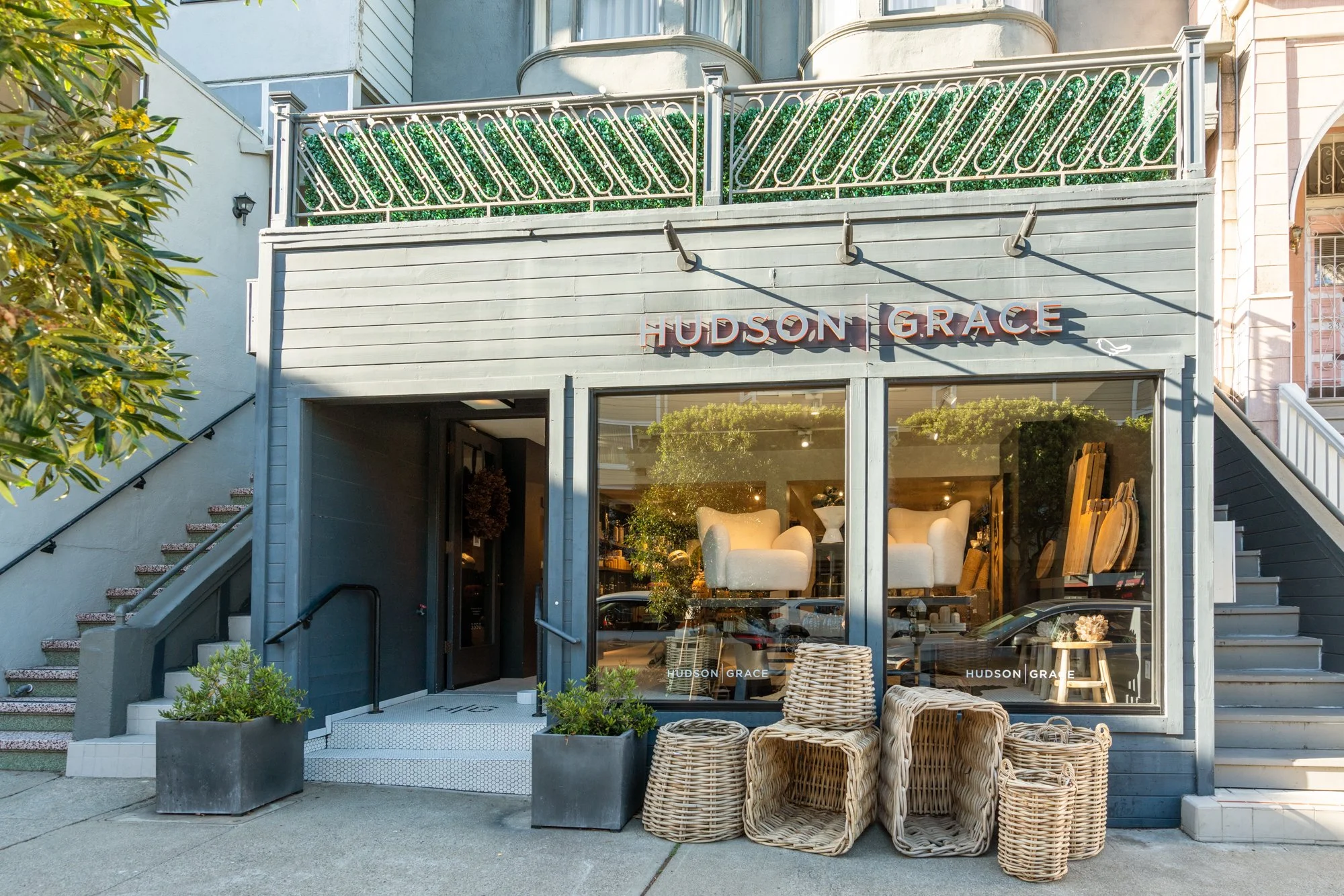 Storefront of Hudson Grace with large front windows displaying furniture inside, outdoor wicker chairs and planters outside, and a sign with the store's name on the building.