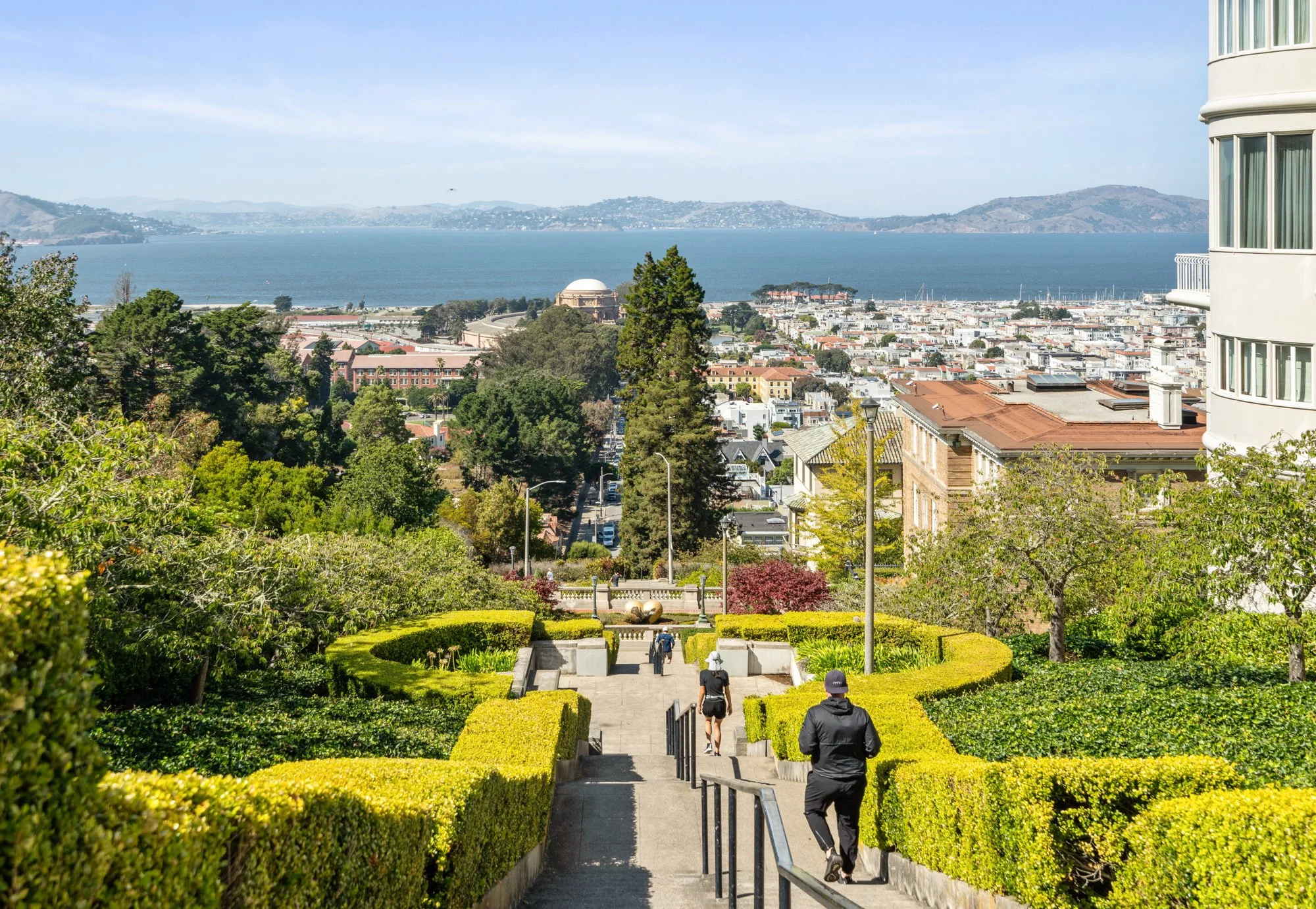 People walking down a sidewalk in a lush green park with city and water in the background, and hills in the distance.