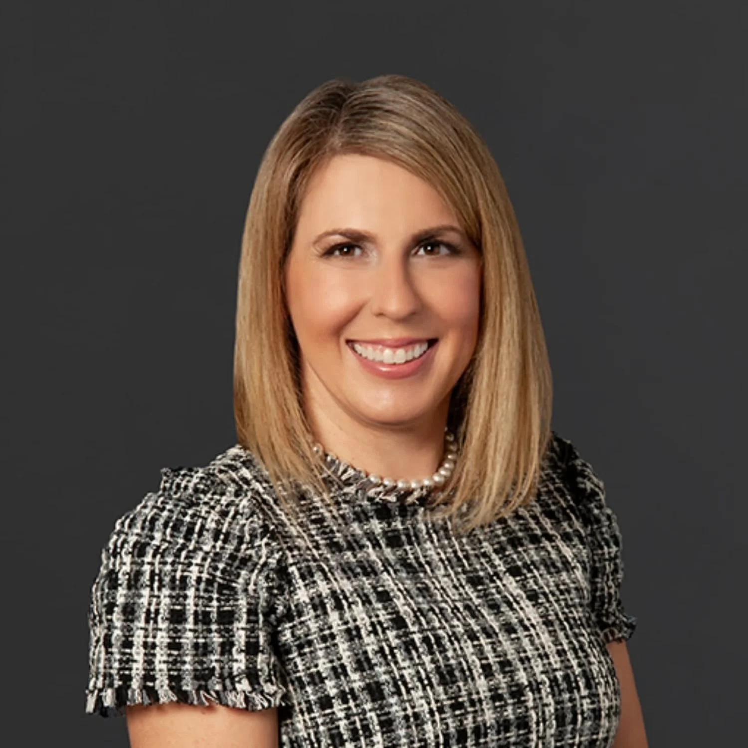 Portrait of a woman with shoulder-length blonde hair, wearing a black and white patterned top and a pearl necklace, smiling against a dark gray background.