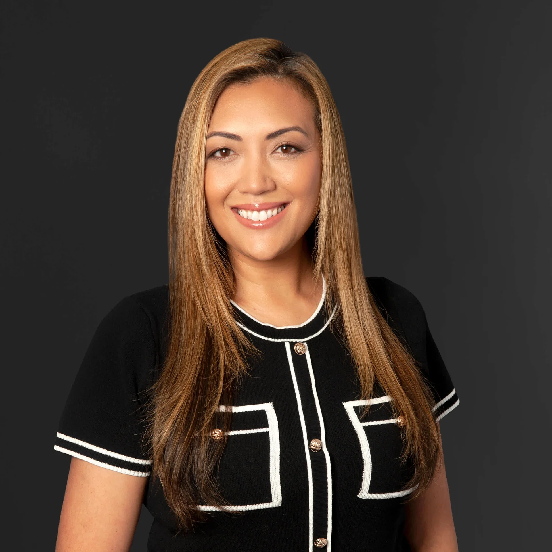 A woman with long, straight hair smiling, wearing a black dress with white piping and gold buttons, standing against a dark background.