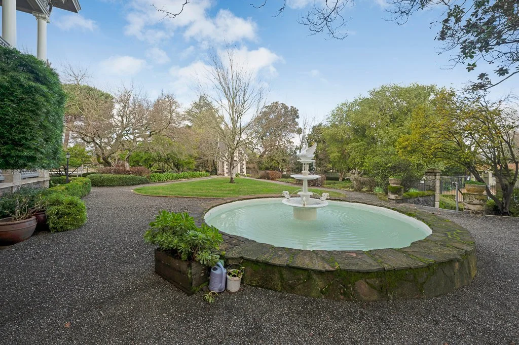A peaceful garden scene with a small fountain in the middle of an oval-shaped pond, surrounded by lush green trees, shrubs, and a paved pathway.