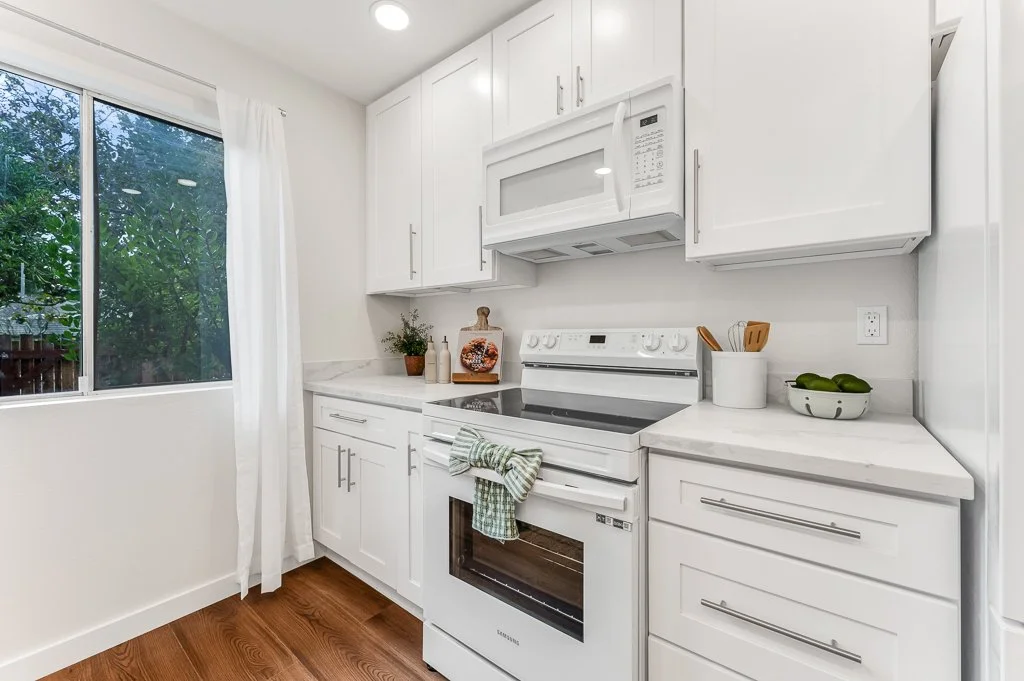 White kitchen with cabinets, oven, microwave, and window with white curtains, showing trees outside.