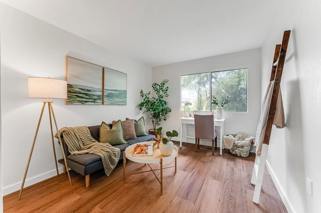 Cozy living room with a gray sofa decorated with pillows and a beige blanket, a round coffee table with a plant, a white desk with a chair near a window, a tall standing lamp, a wall art piece with ocean scenery, and a ladder leaning against the wall