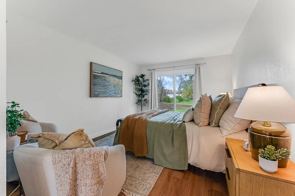 A cozy bedroom with a bed against a window, a beige armchair with cushions, a side table with a lamp and small potted plants, and a painting of a beach on the wall.