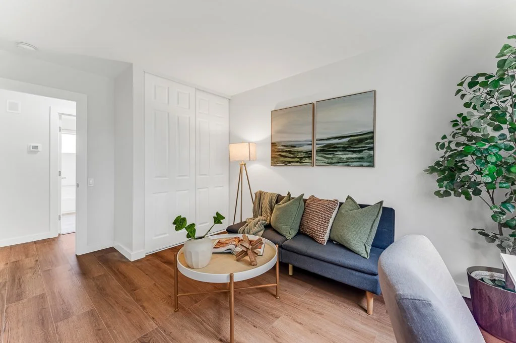 Living room with navy blue sofa, multiple pillows, a floor lamp, wall art of two landscape paintings, a coffee table with plants and decorative items, and wooden flooring.