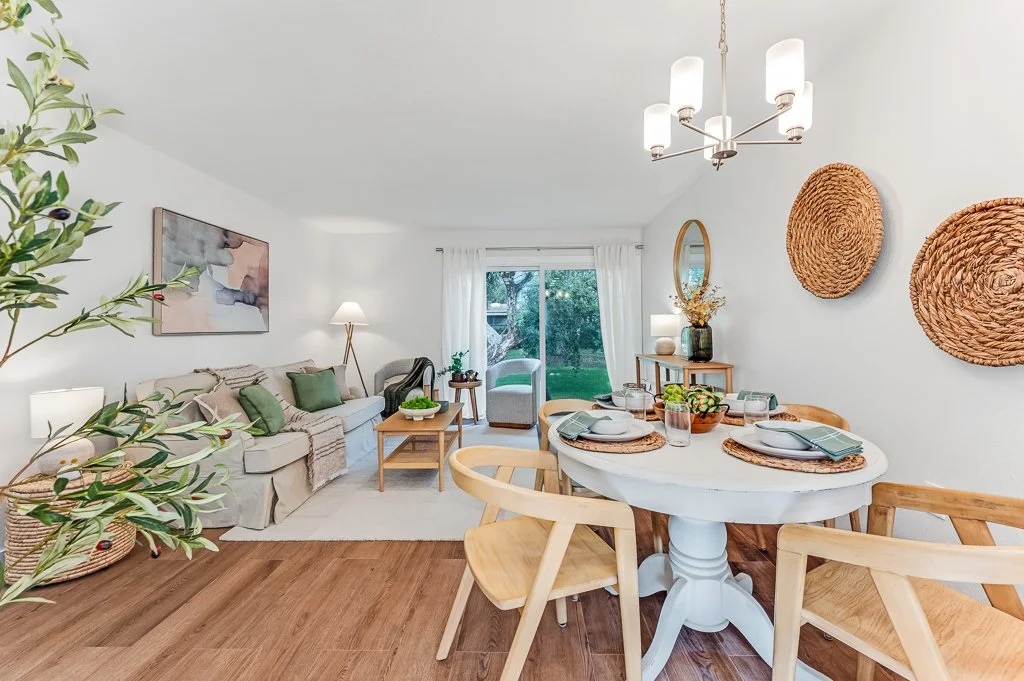 Living room and dining area with a white sofa, green pillows, wall art, and a wooden dining table with four chairs, dishes, and decorative items, with a sliding glass door leading outside.