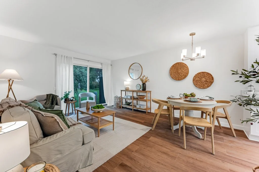Living and dining room with beige sofa, white curtains, wooden coffee table, green plants, round mirror, wooden dining table with six chairs, wall art, and a chandelier.