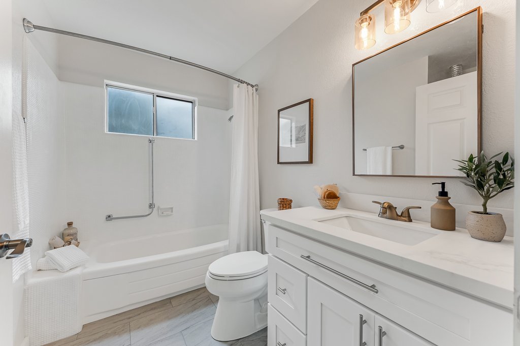 Bright bathroom with white vanity, marble countertop, a large mirror, and a bathtub with a frosted window above. Decor includes a plant, soap dispenser, and framed pictures.