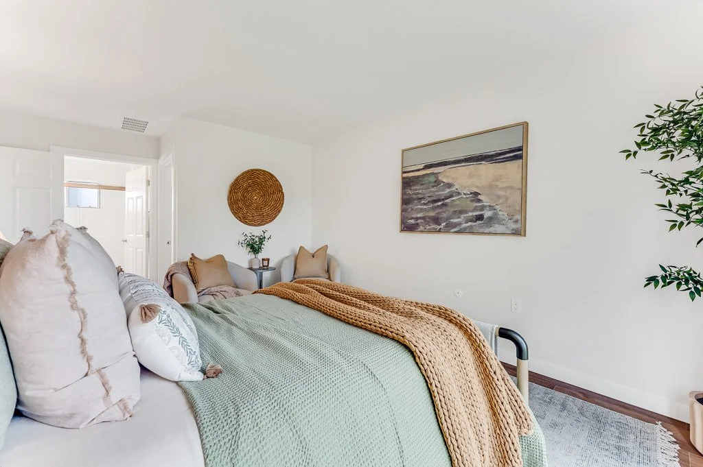 A cozy bedroom with a bed covered in a green blanket and brown throw, two beige pillows, and decorative pillows. There are two armchairs with pillows and a side table with a plant. A framed painting of a beach scene hangs on the wall, and a large lea