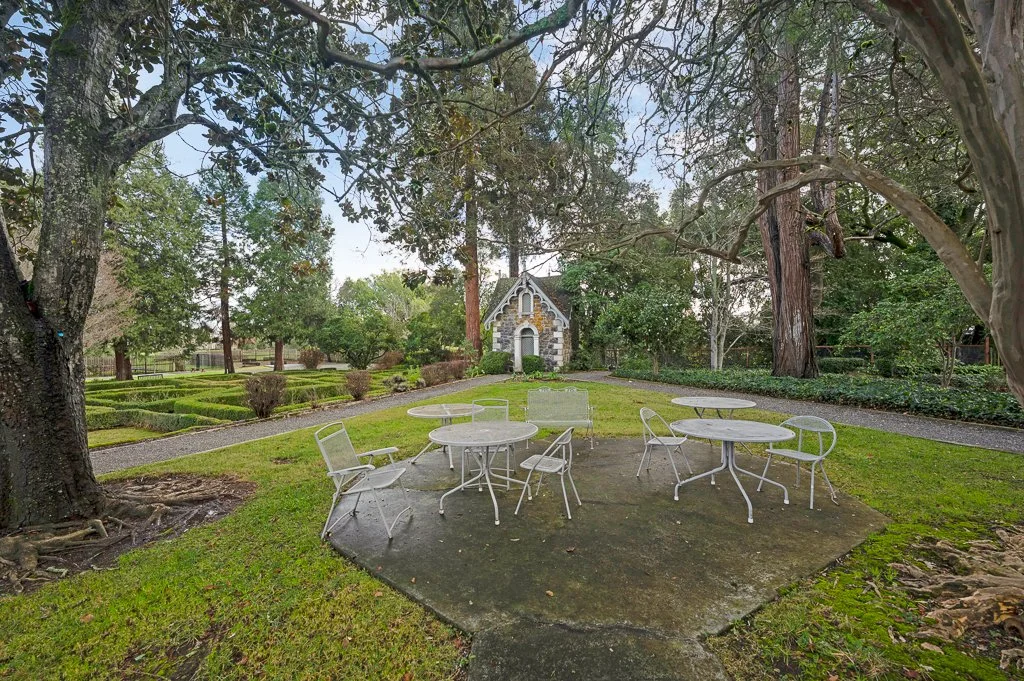 Outdoor garden with white metal patio tables and chairs on a concrete pad, surrounded by trees, bushes, and landscaped gardens, with a small stone building in the background.