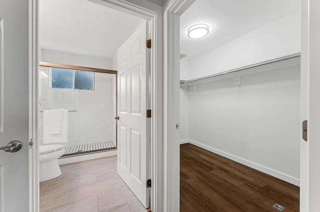 An open doorway reveals a bathroom on the left with a toilet, tiled shower area, a small window, and white walls; the right side shows an empty walk-in closet with white walls, a wooden floor, and built-in shelving.