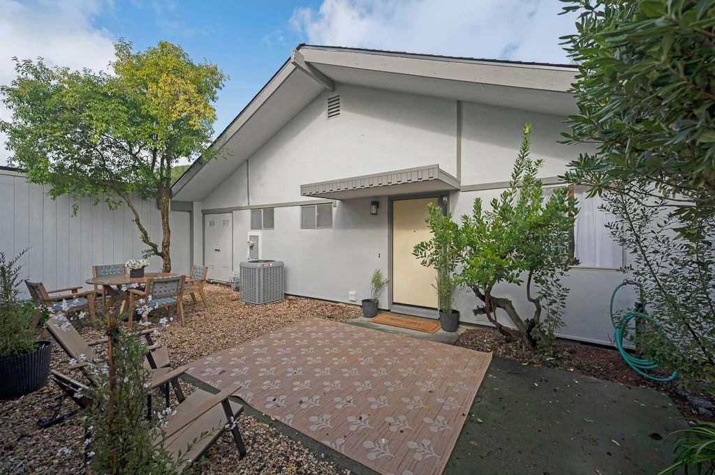 Backyard patio area with a tan rug, outdoor furniture including a table with chairs, and trees along the house exterior with a white wall, door, and windows.