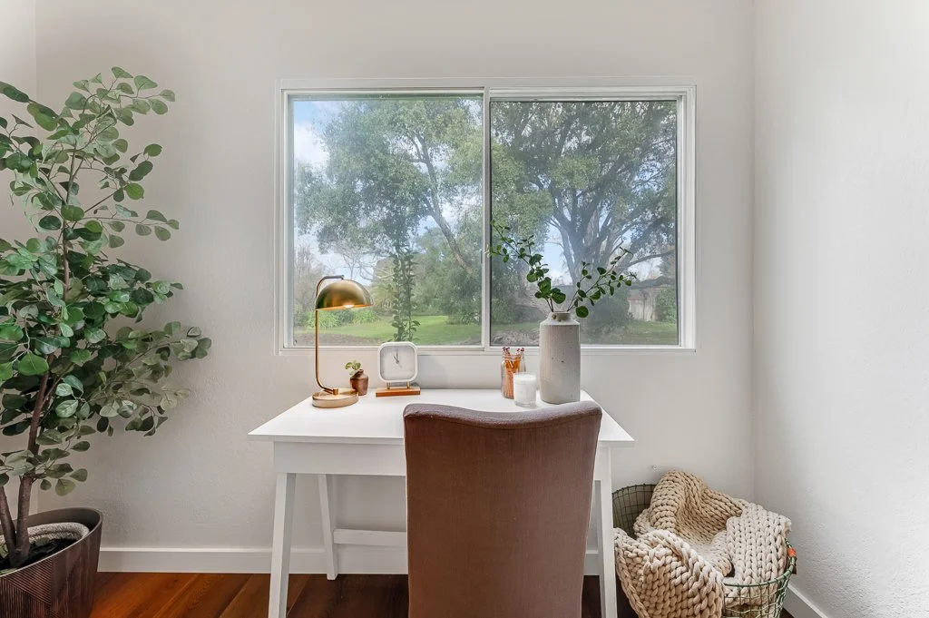 Home workspace with white desk, brown upholstered chair, large window overlooking trees, potted plant, gold desk lamp, clock, and decorative items, with a cozy blanket in a basket beside the desk.