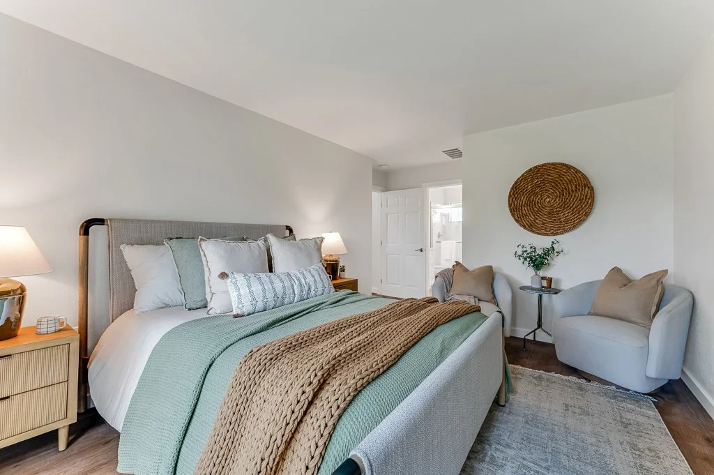 Bedroom with a light-colored upholstered bed, multiple pillows, a green blanket, and a chunky brown throw. Nightstands with lamps on either side. Two beige armchairs with pillows and a small table with a plant and a woven wall decor are against the w