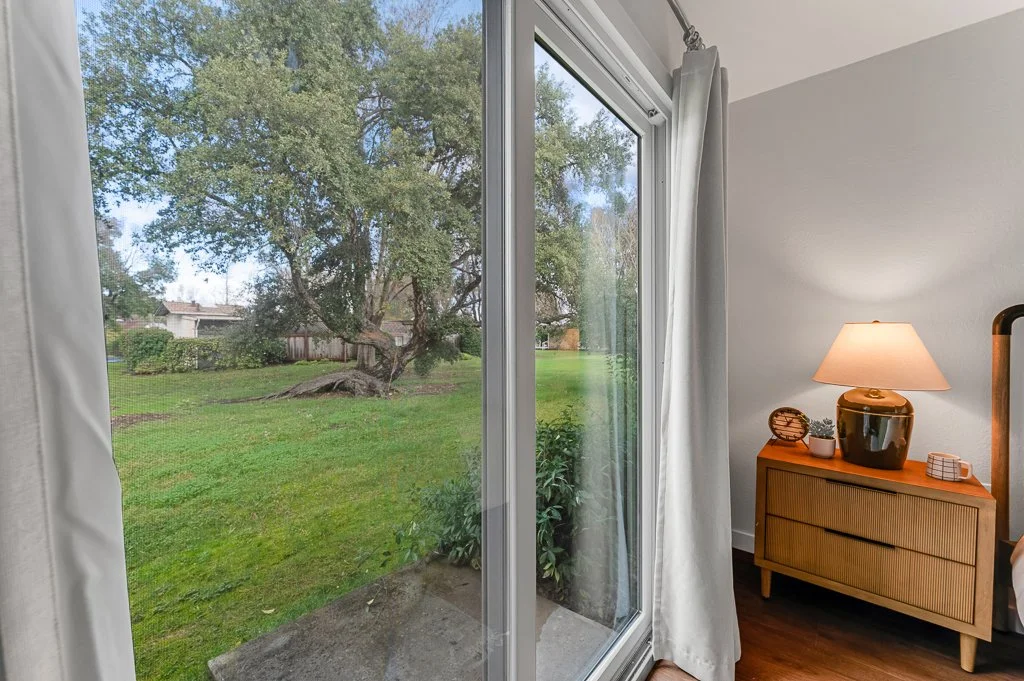 View of a backyard with green grass and trees through a sliding glass door, with part of an indoor side table with a lamp on it visible.