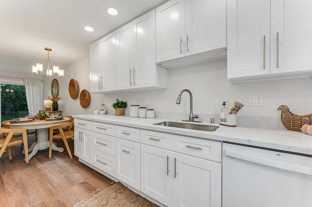 Bright kitchen with white cabinets, marble countertop, and stainless steel faucet, adjacent to a dining area with a round wooden table and four chairs, hanging light fixture, and decorative wall hangings.