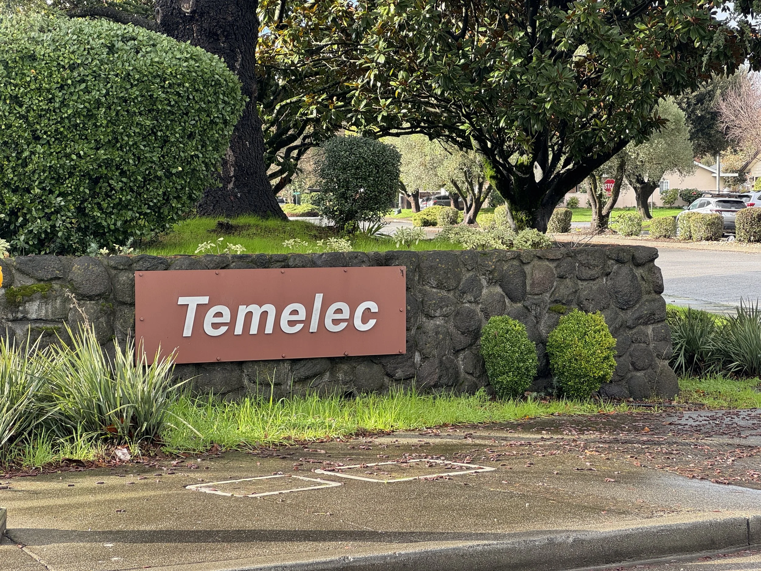 A stone wall with a pink sign reading 'Temelec' in front of a lush landscape with grass, bushes, and trees, near a street with parked cars.